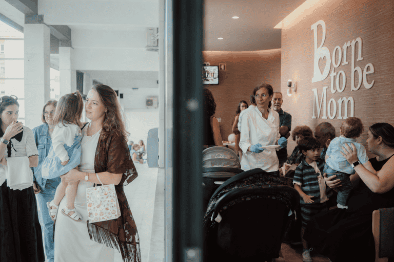 A woman carries a child and a gift bag as she walks through a bright hospital hallway toward a maternity waiting area with people gathered inside.