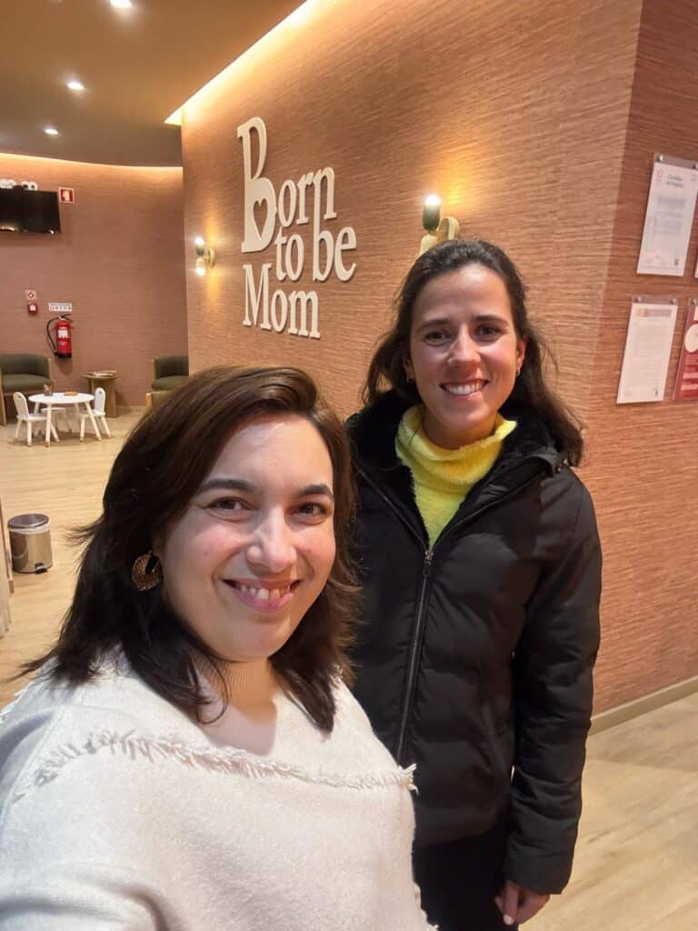 Two women smiling for a selfie in a warm lobby with a large 'Born to be Mom' sign on a wood-paneled wall behind them