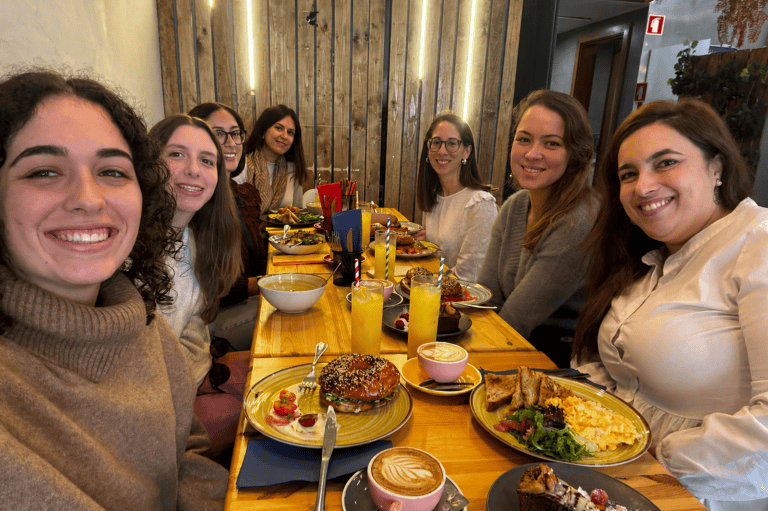 Eight friends smiling and eating at a long wooden table with plates of brunch, coffee, and orange drinks in a casual restaurant setting.