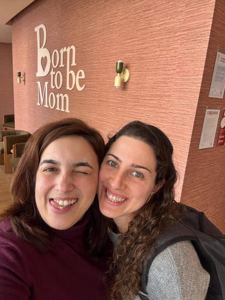 Two women smiling for a selfie in a hallway with a pink textured wall and the words 'Born to be Mom' visible behind them.